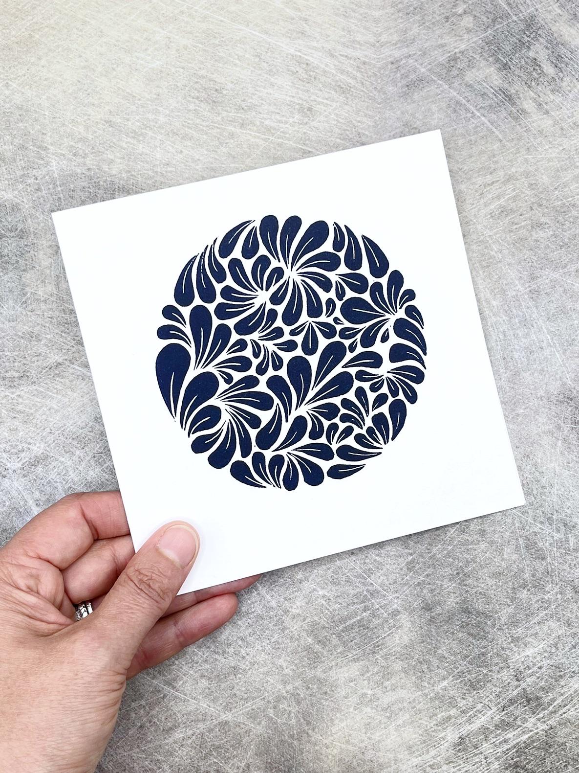Hand holding a white card with a lino printed blue mussel shell pattern laid on a grey surface
