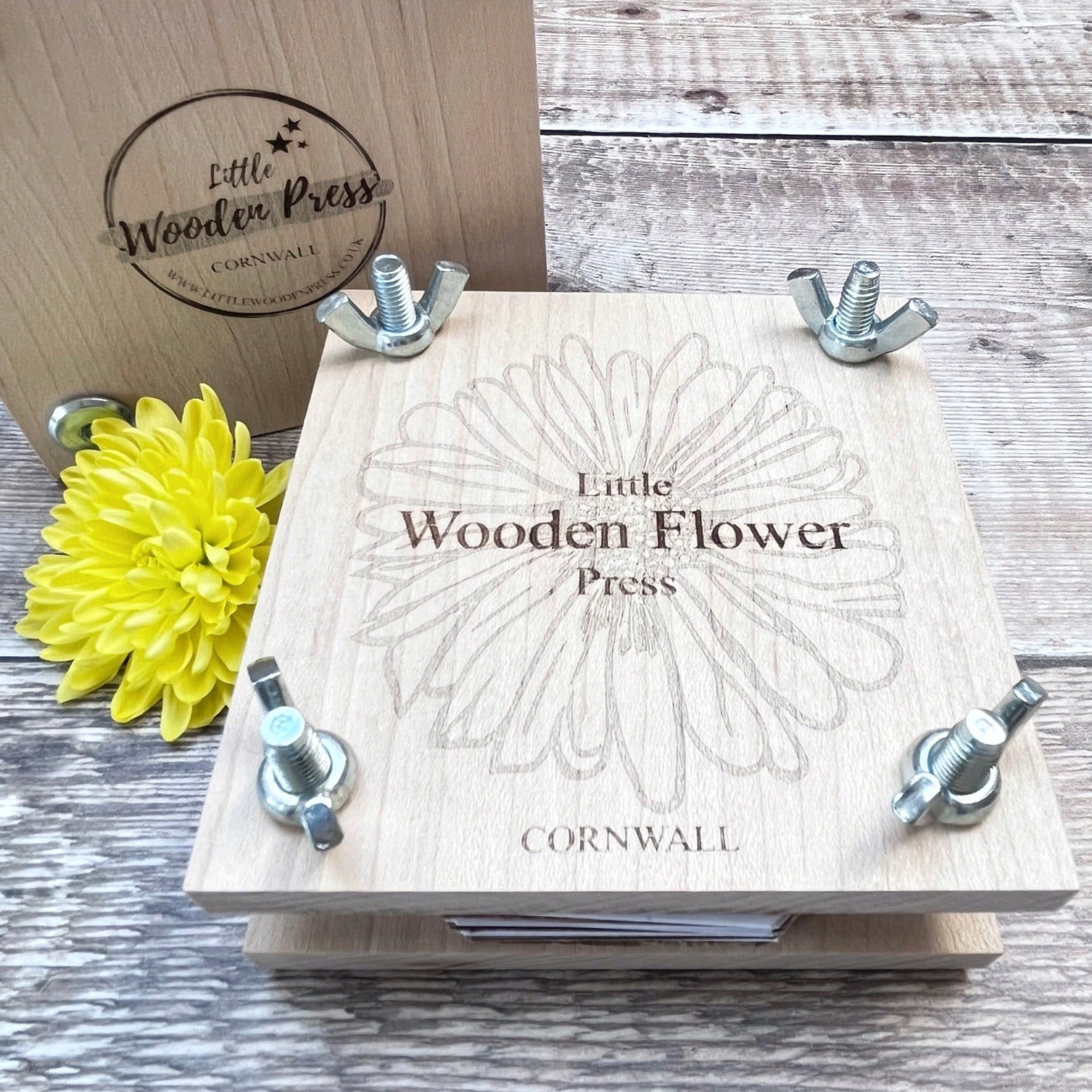 Wooden flower press with a yellow flower and branded lid laid on a wooden surface