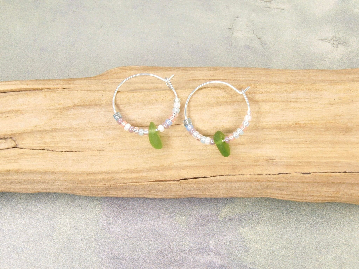 Silver hoop earrings with pastel seed beads and green sea glass laid on a piece of driftwood.