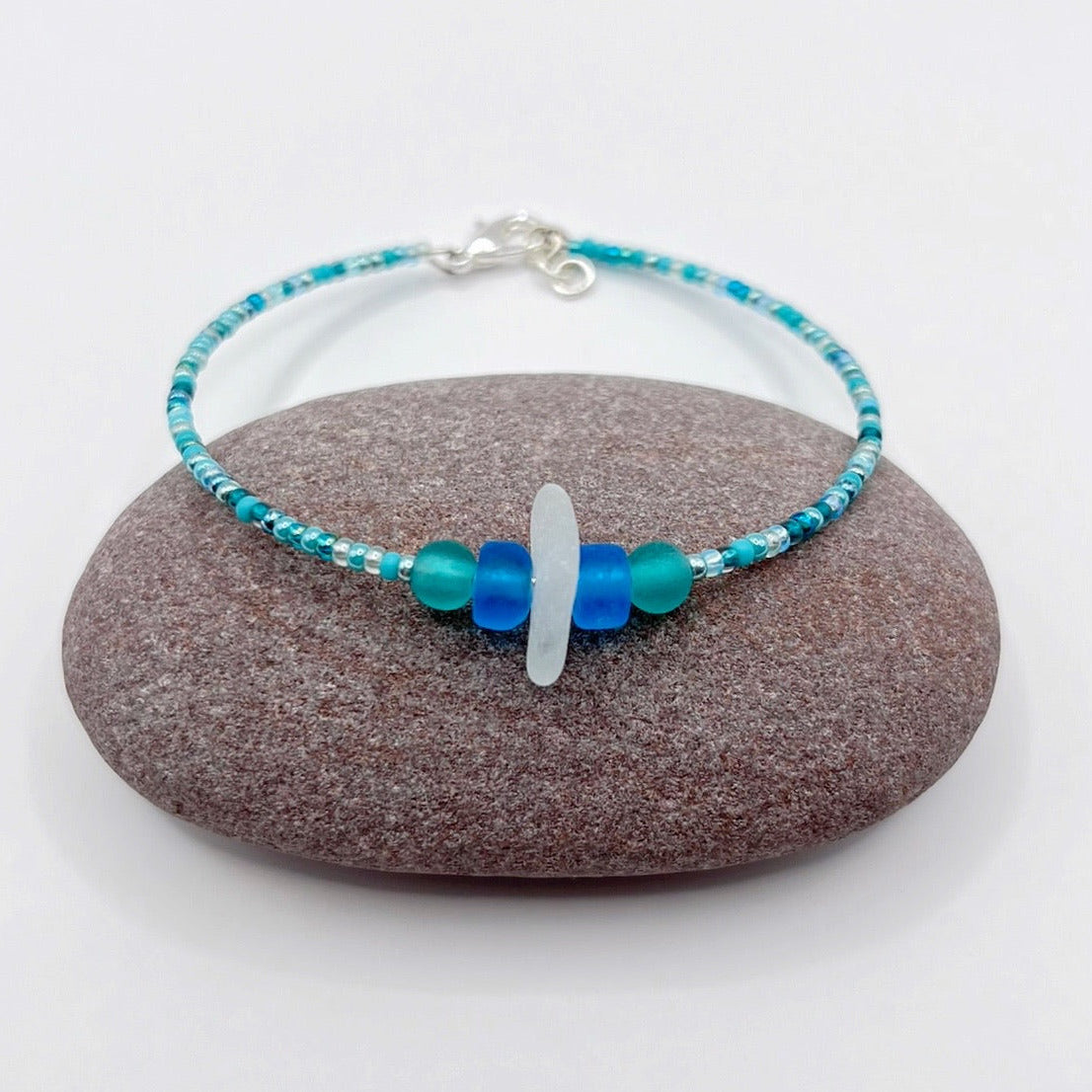 Bracelet with white sea glass and blue and turquoise beads laid on a purple pebble against a white background
