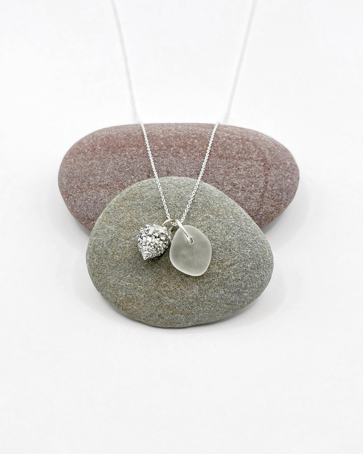 Silver necklace with White Sea glass and silver shell pendant laid across two pebbles against a white background