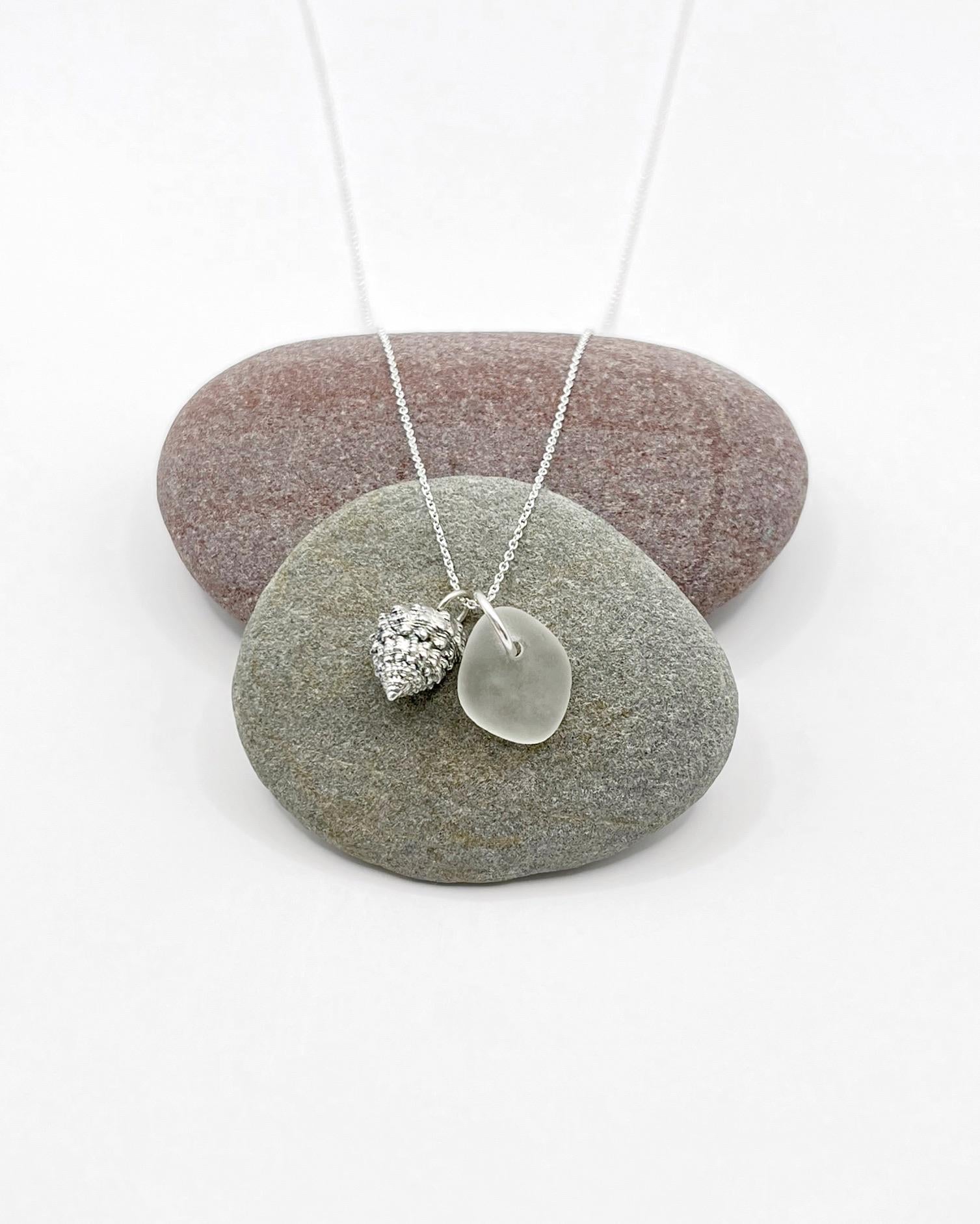 Silver necklace with White Sea glass and silver shell pendant laid across two pebbles against a white background