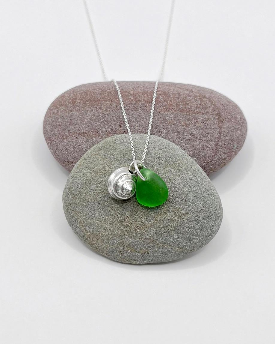 Necklace with a silver shell and green sea glass laid on a grey pebble against a white background