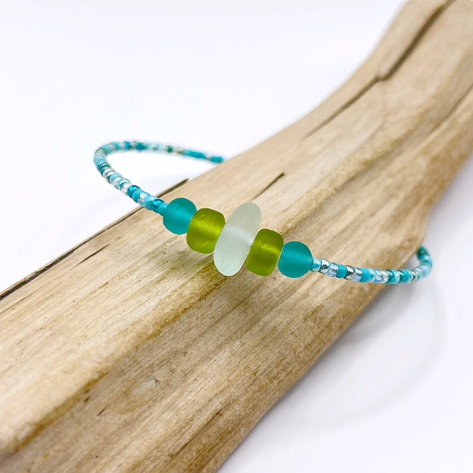 A turquoise beaded bracelet with lime and sea green frosted glass beads and a piece of sea foam coloured Cornish sea glass, finished with a silver plated lobster clasp and laid on a piece of driftwood.