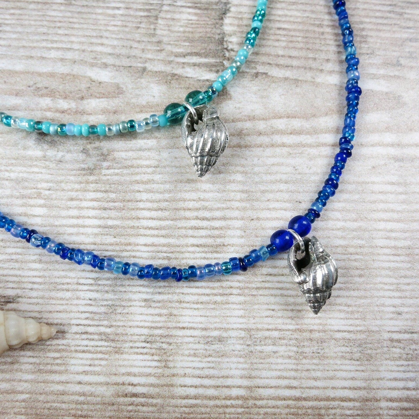 Two blue seed beaded necklaces with silver shell pendants laid on a textured surface.