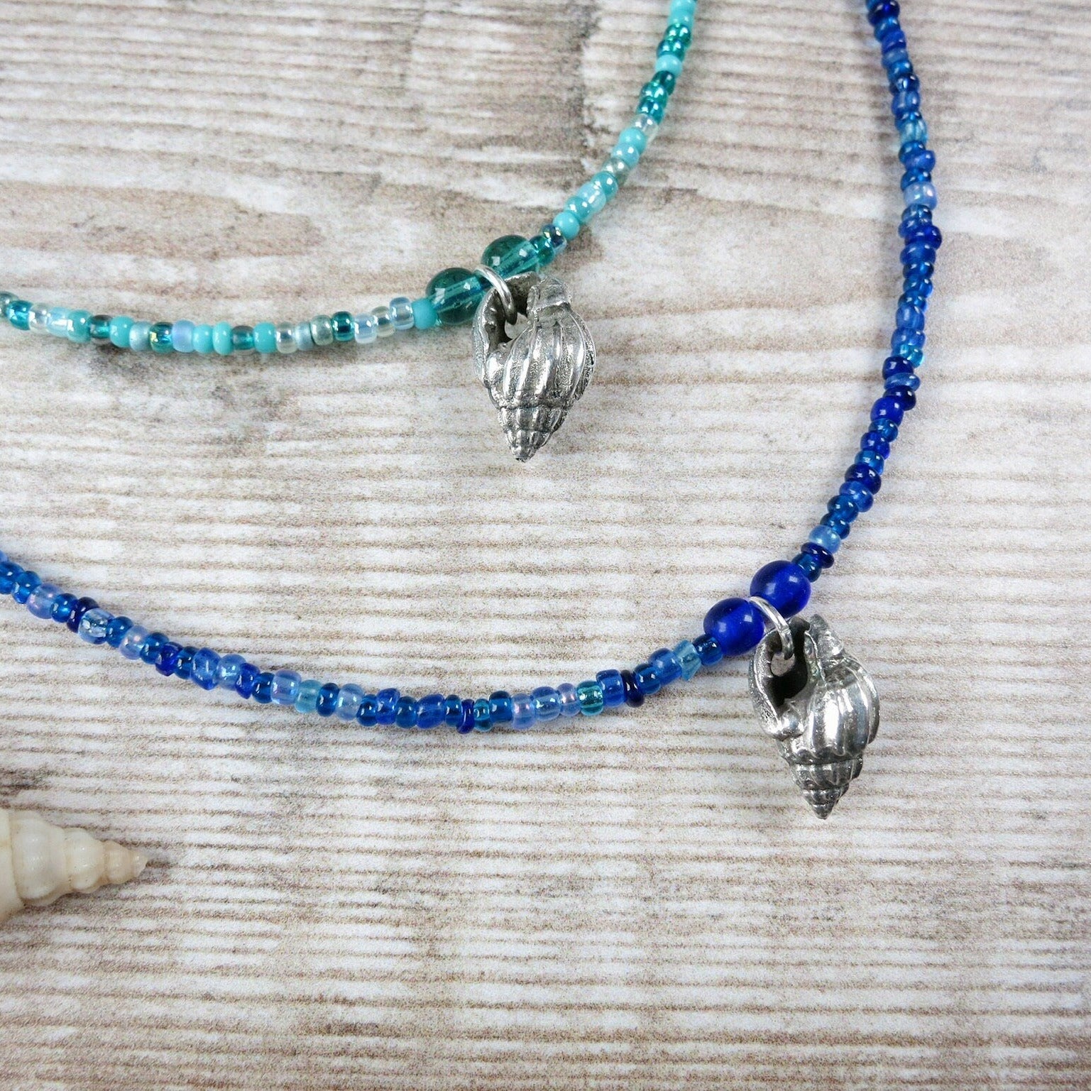 Two blue seed beaded necklaces with silver shell pendants laid on a textured surface.