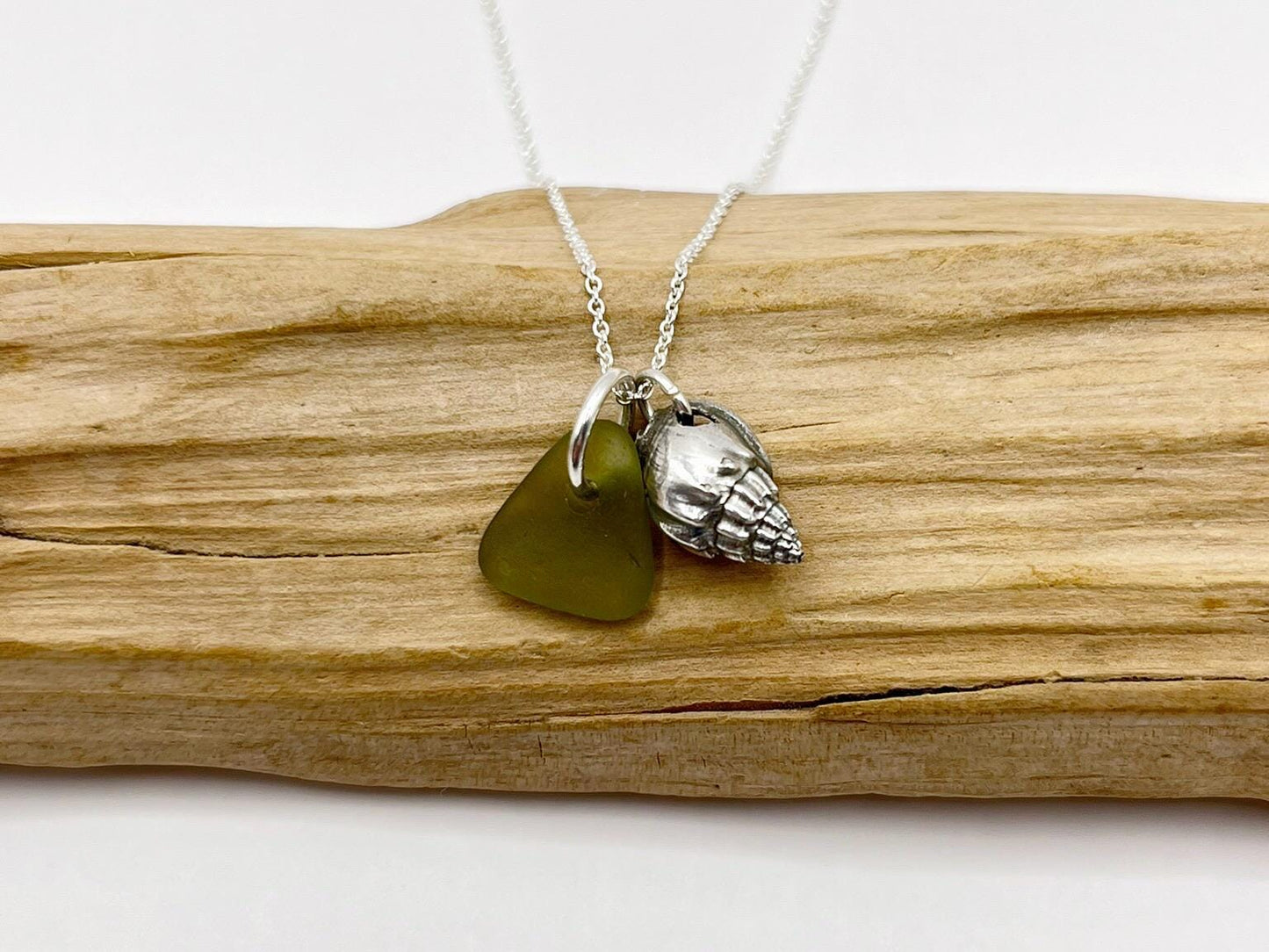 Necklace with olive green sea glass and a silver shell charm laid on driftwood