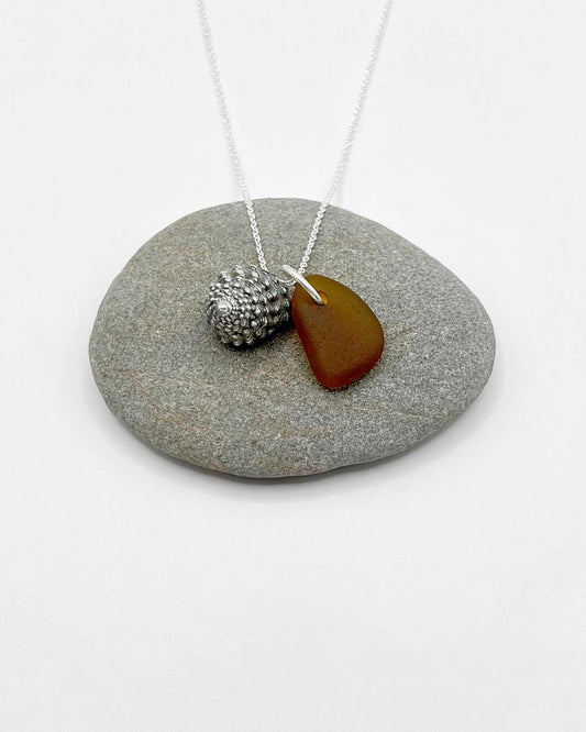 Necklace with burnt orange sea glass and silver shell pendant laid on a grey pebble against a white background
