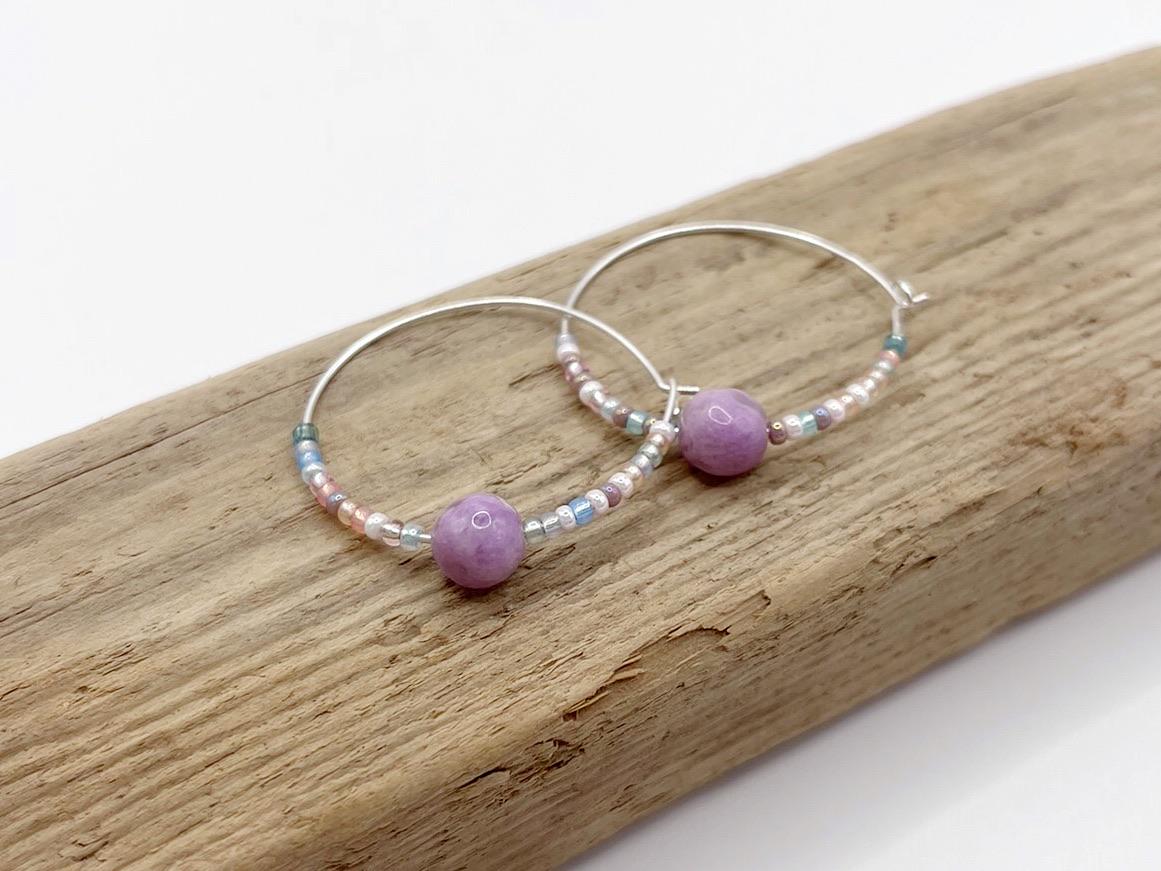 Close up of a pair of silver hoop earrings with lilac Jasper and seed beads laid on driftwood against a light surface.