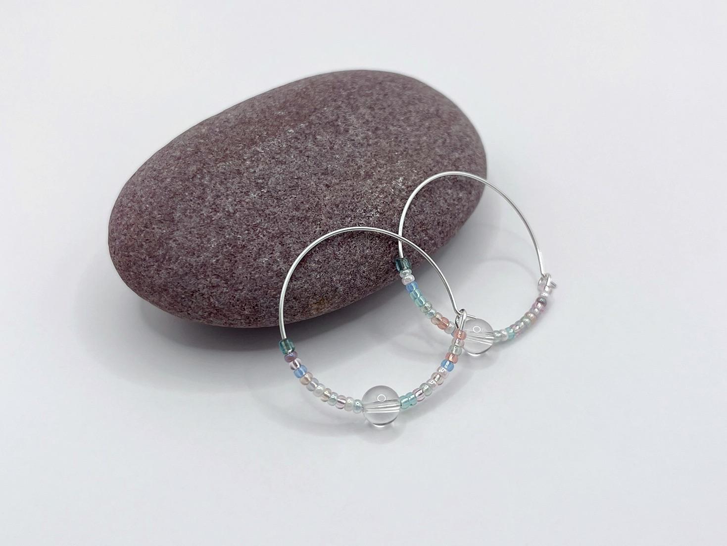 Pair of pastel seed bead and clear crystal hoop earrings laid against a purple pebble on a white surface