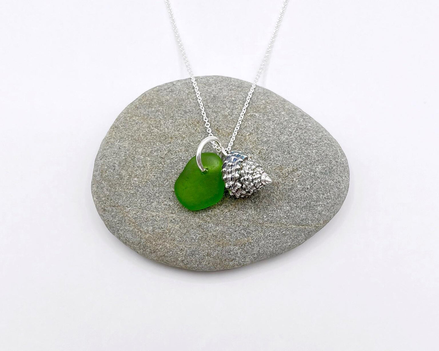 Necklace with green sea glass and silver shell pendant laid on a grey pebble against a white background