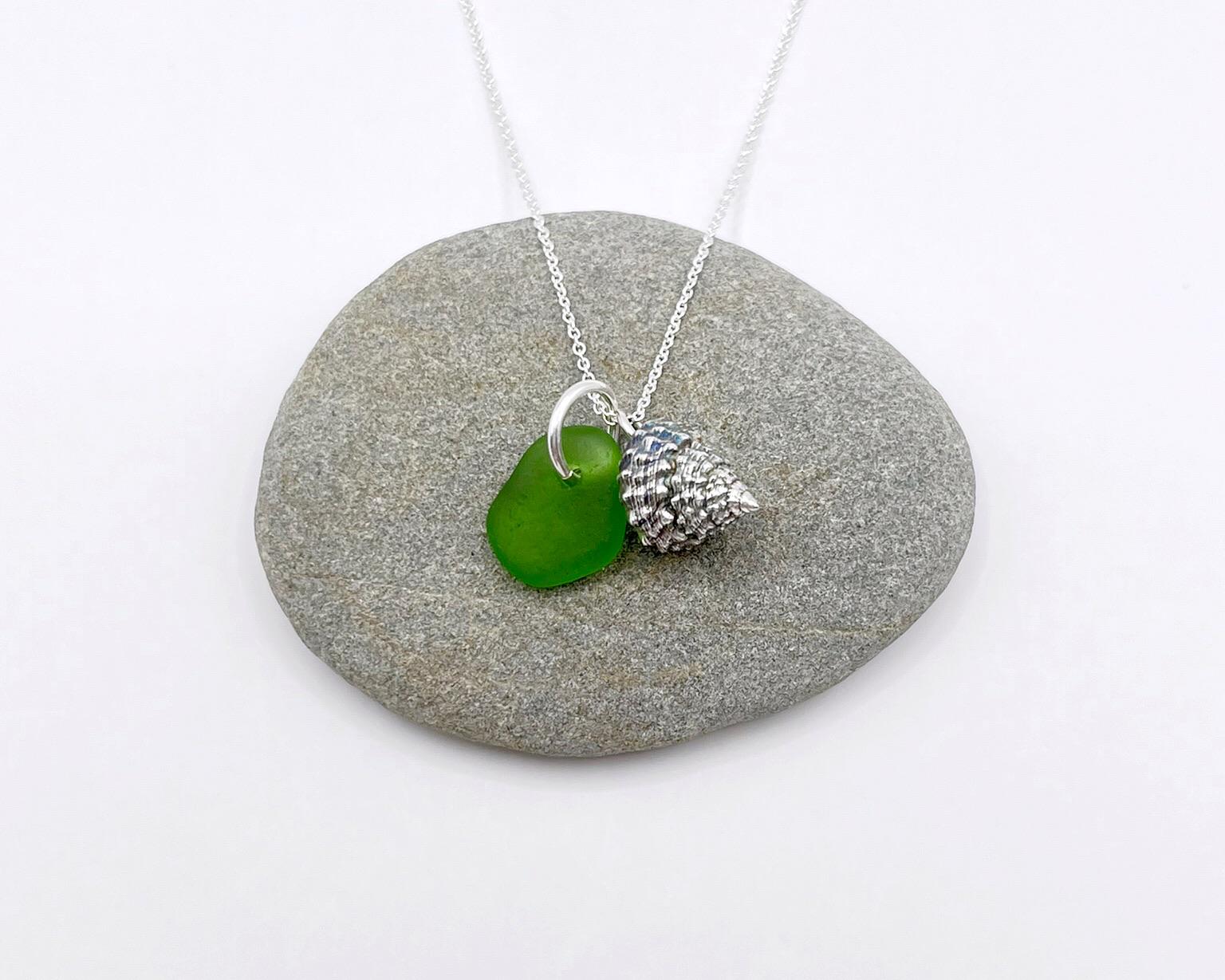 Necklace with green sea glass and silver shell pendant laid on a grey pebble against a white background
