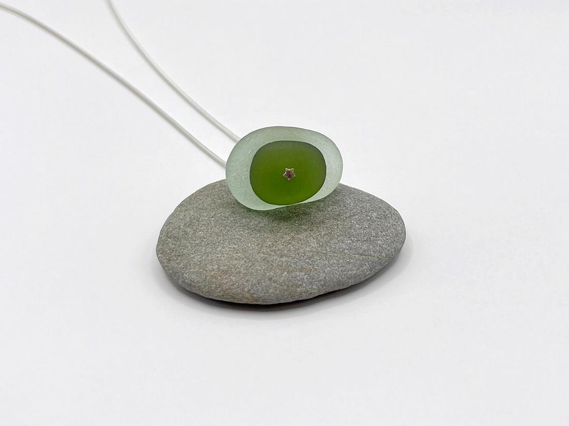 Close up showing the silver star at the base of the sea glass pendant, laid on a pebble against a white background 
