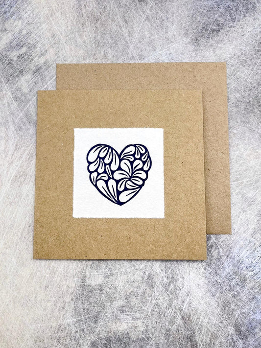 Kraft card with a white square featuring an original lino printed blue heart design with matching Kraft envelope, laid on a textured grey background.