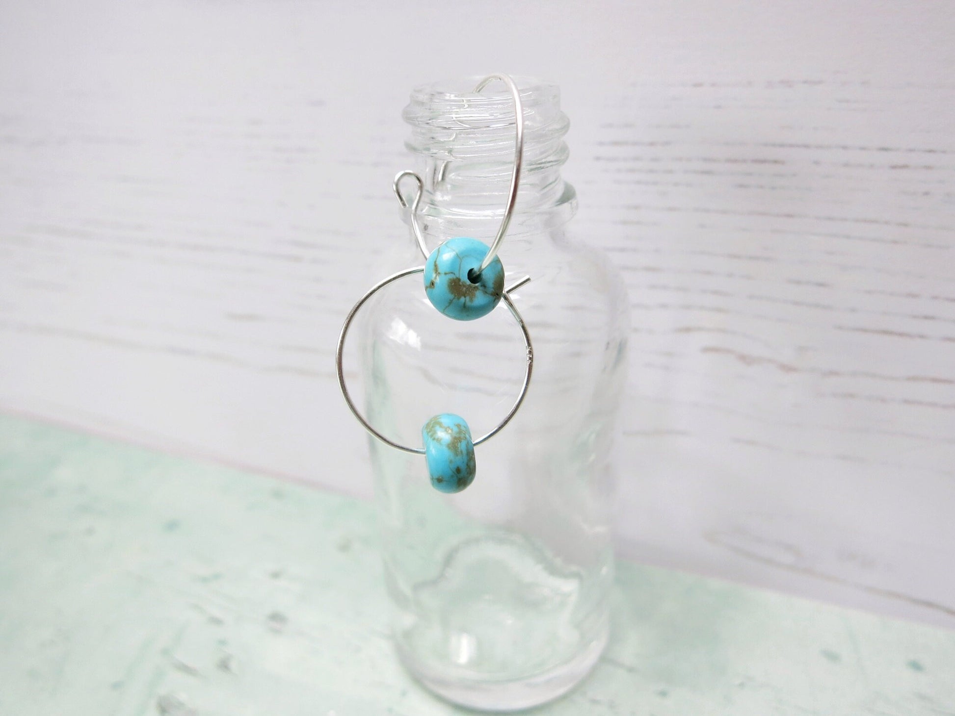 Silver hoop earrings with turquoise beads hanging on a glass bottle against a pale background