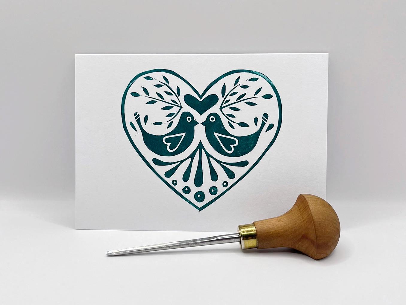 White card with an original lino printed green heart design featuring two birds stood against a white background. Photo includes lino cutting tool.