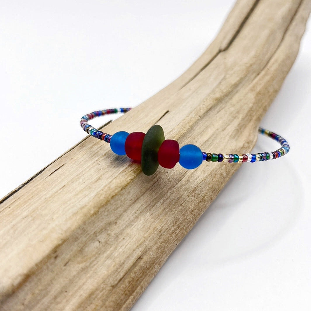 A rainbow coloured beaded bracelet with red and blue frosted glass beads and a piece of olive green coloured Cornish sea glass, finished with a silver plated lobster clasp and laid on a piece of driftwood.