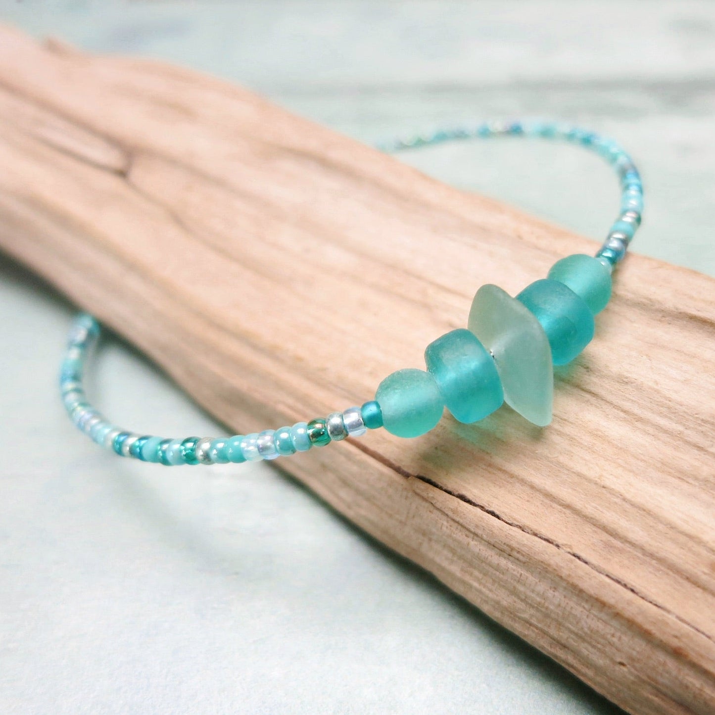 Close up of bracelet with pale sea foam sea glass and sea green and turquoise glass beads laid on driftwood against a pale blue background