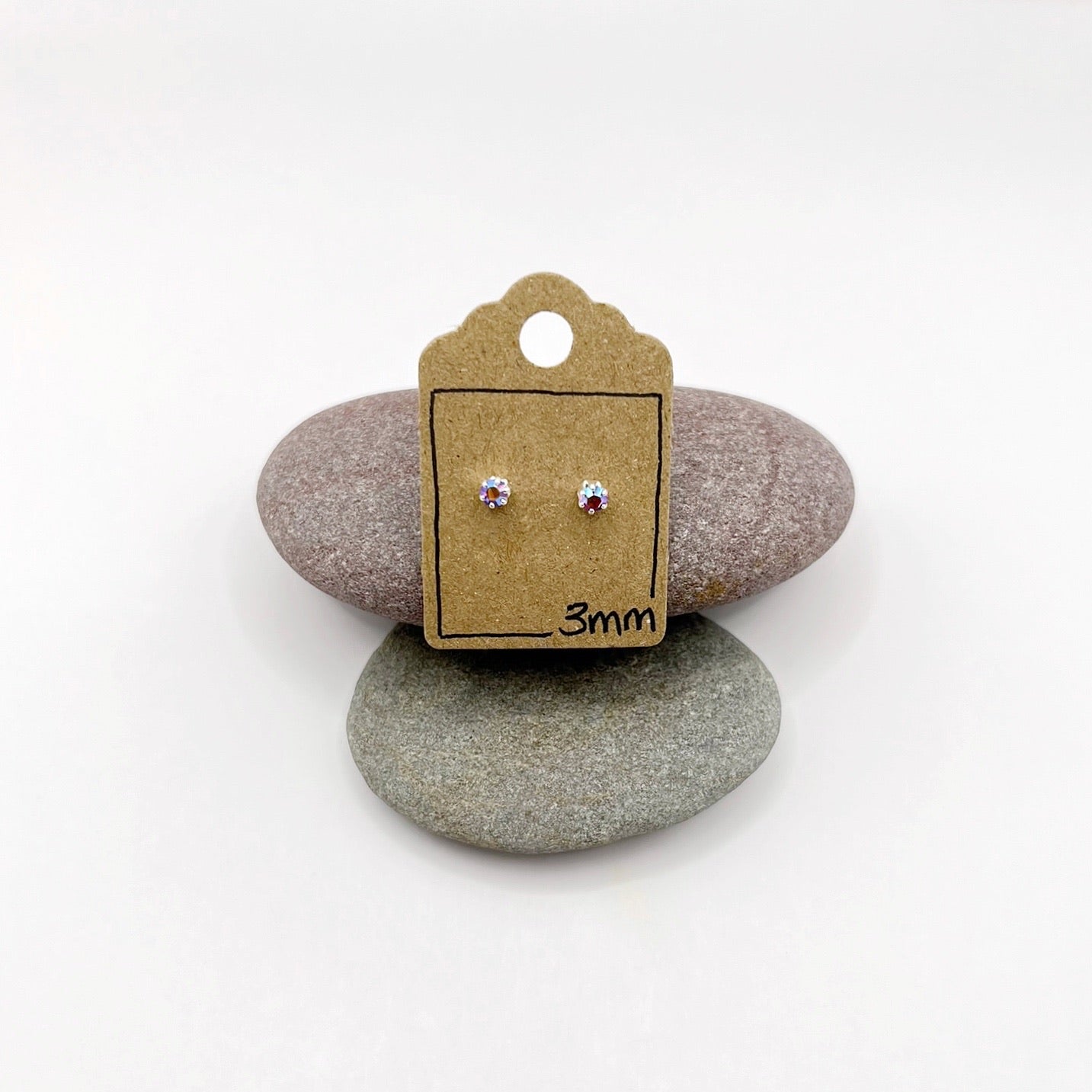Pair of silver stud earrings on a cardboard backing card with '3mm' marking, placed on two stones.