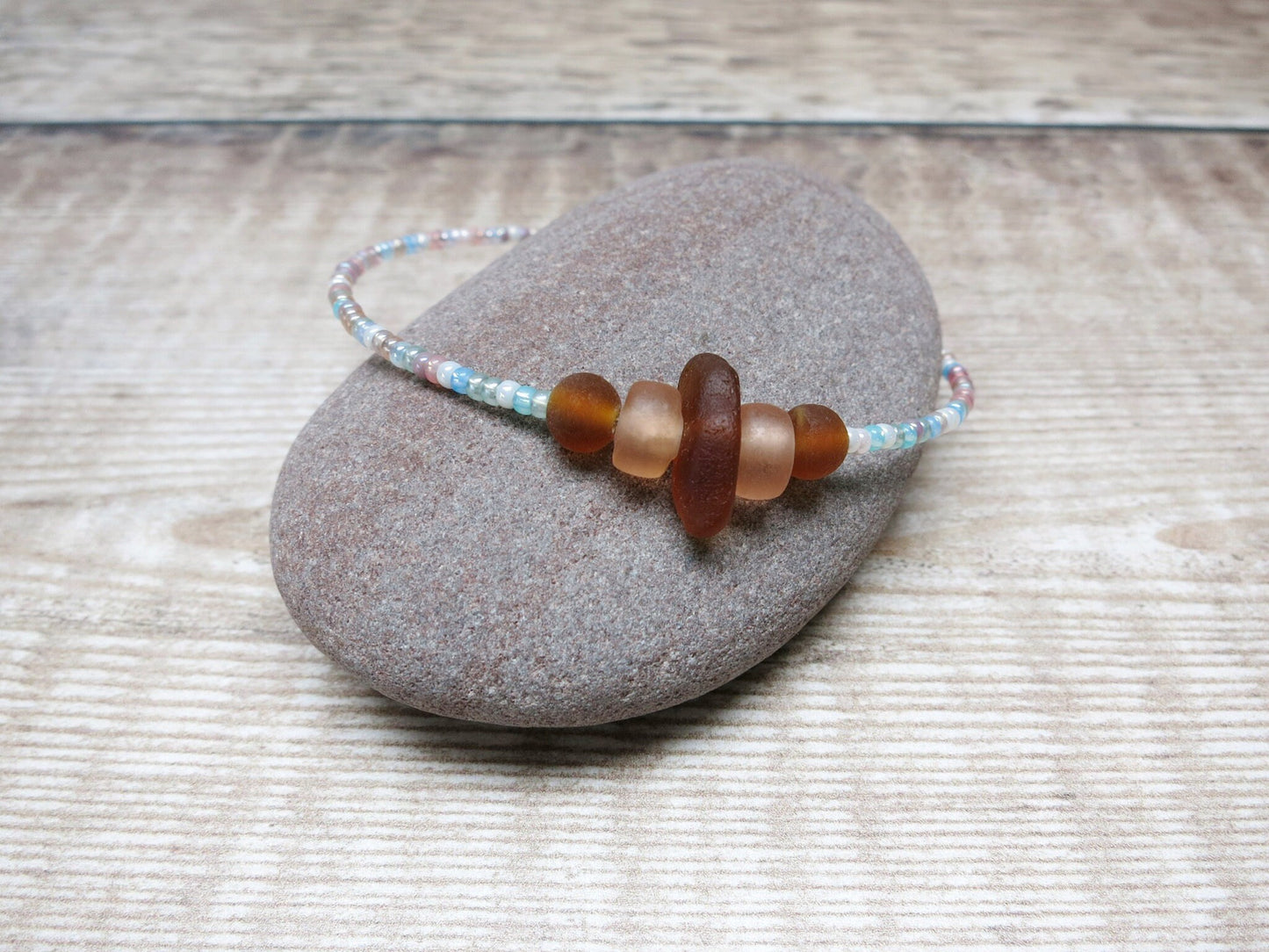 Bracelet with pastel seed beads and burnt orange sea glass laid on a pebble against a textured background.