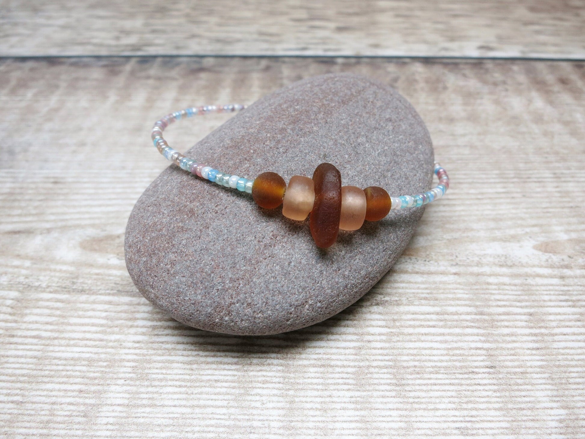 Bracelet with pastel seed beads and burnt orange sea glass laid on a pebble against a textured background.