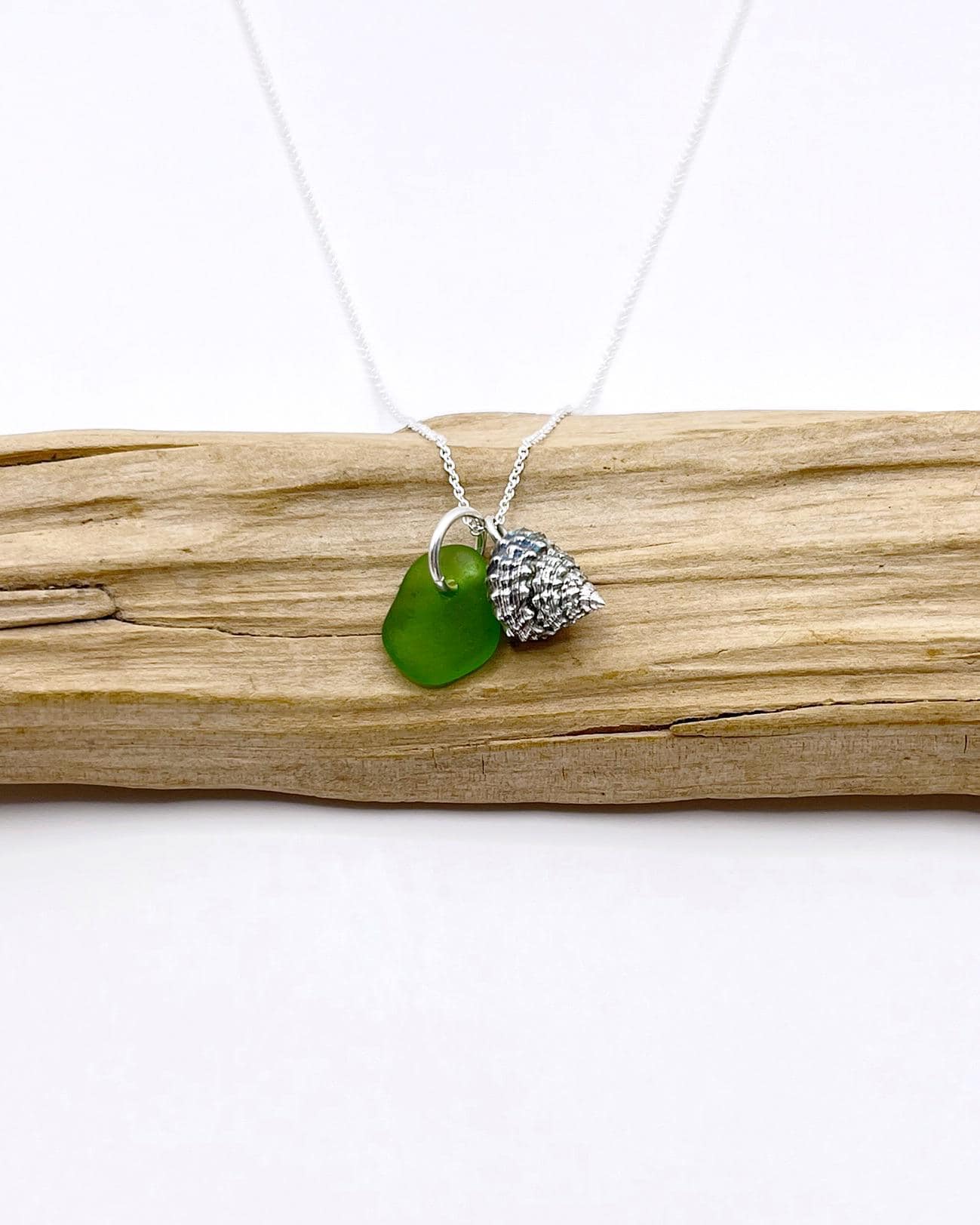 Necklace with green sea glass and silver shell pendant laid on driftwood against a white background
