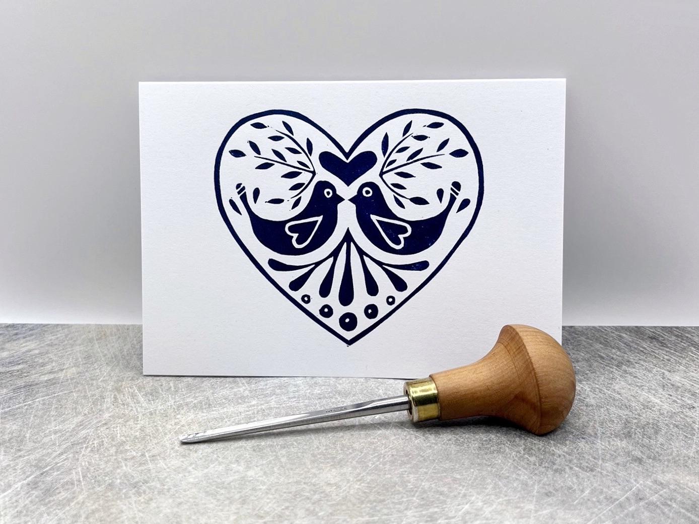 White card with an original lino printed blue heart design featuring two birds. Photo includes lino cutting tool used, stood against a textured background.