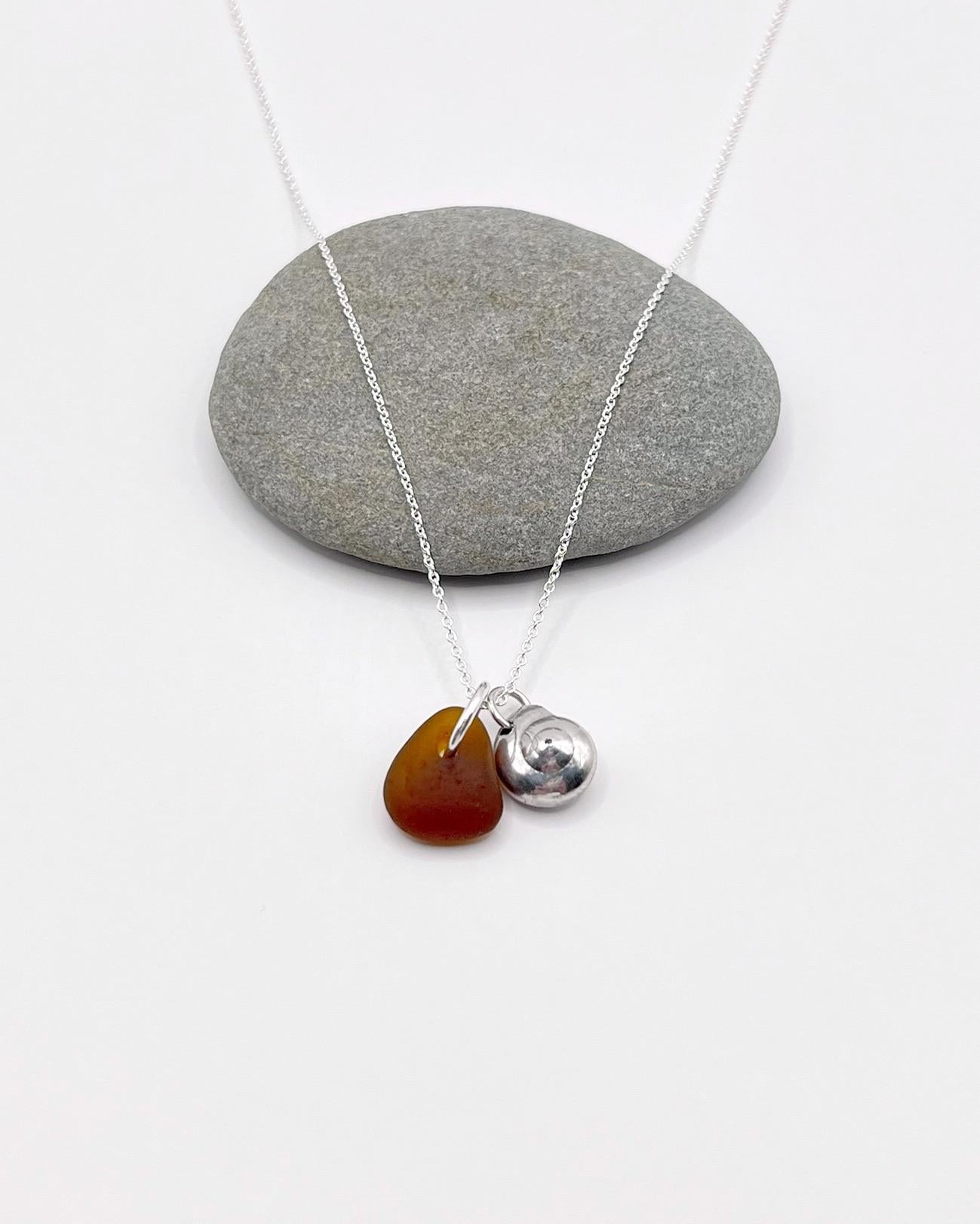 Necklace with burnt orange sea glass and a silver shell pendant, laid in front of a grey pebble on a white surface