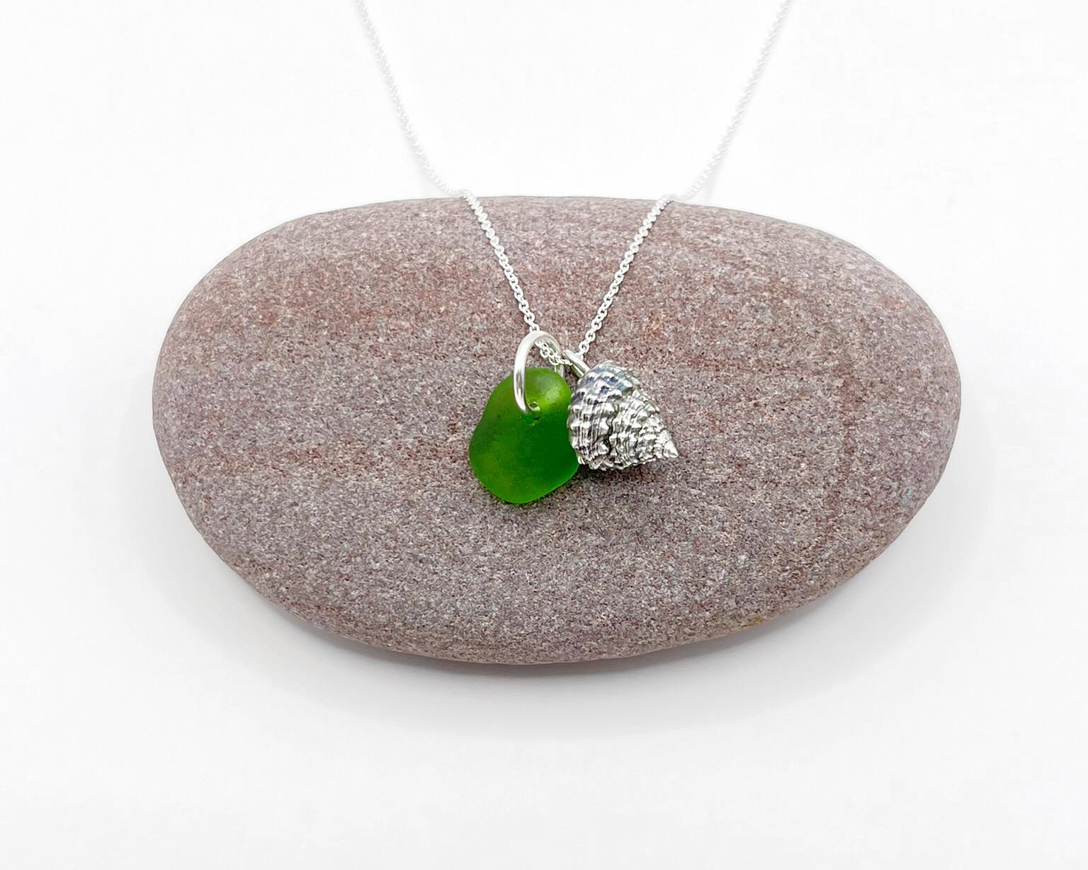Necklace with green sea glass and silver shell pendant laid on a purple pebble against a white background