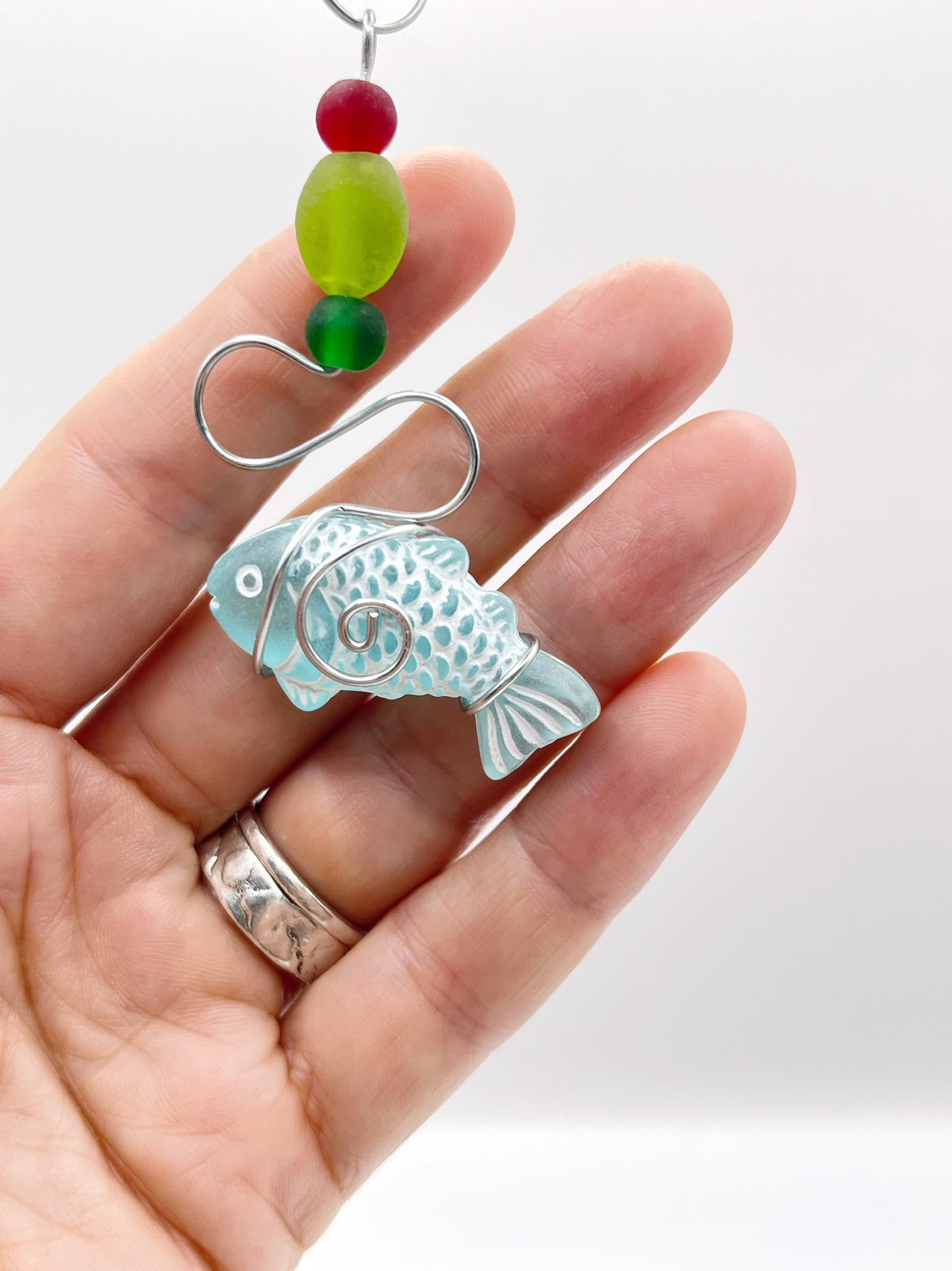 Decorative hanging ornament with a fish design, wire and colourful beads held in a hand against a white background