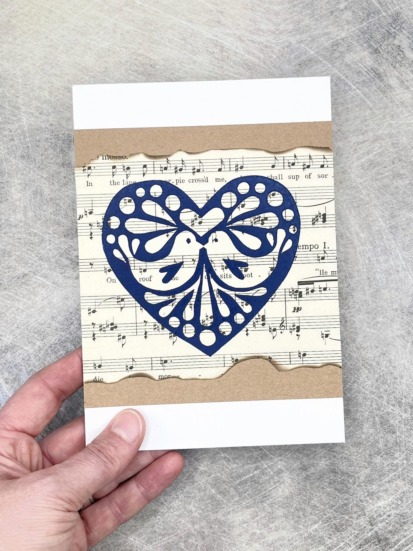 Card with blue heart design on sheet music background held by a hand against a grey textured background