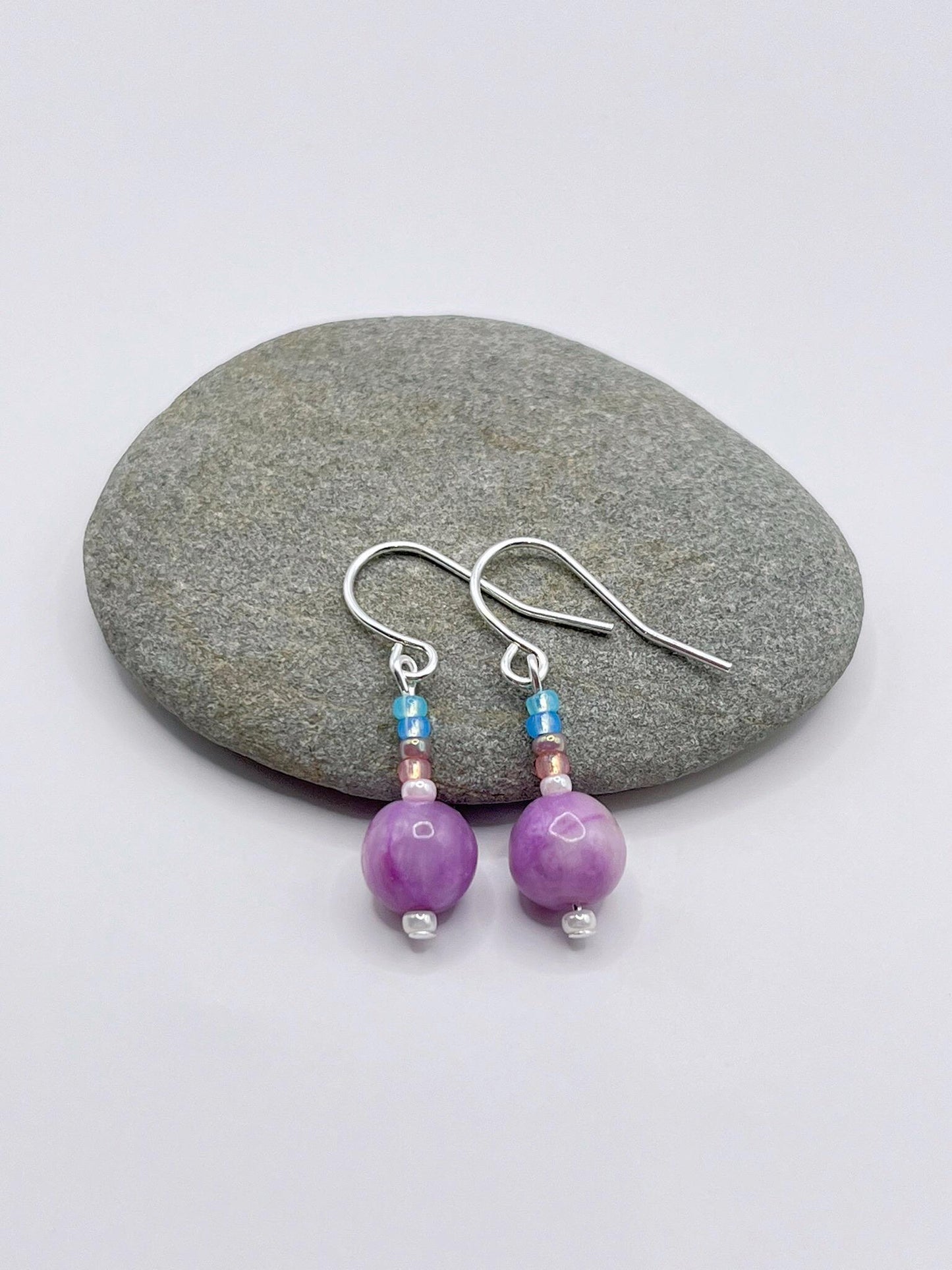 Pair of pastel seed bead and lilac jasper earrings laid on a grey pebble against a white background.