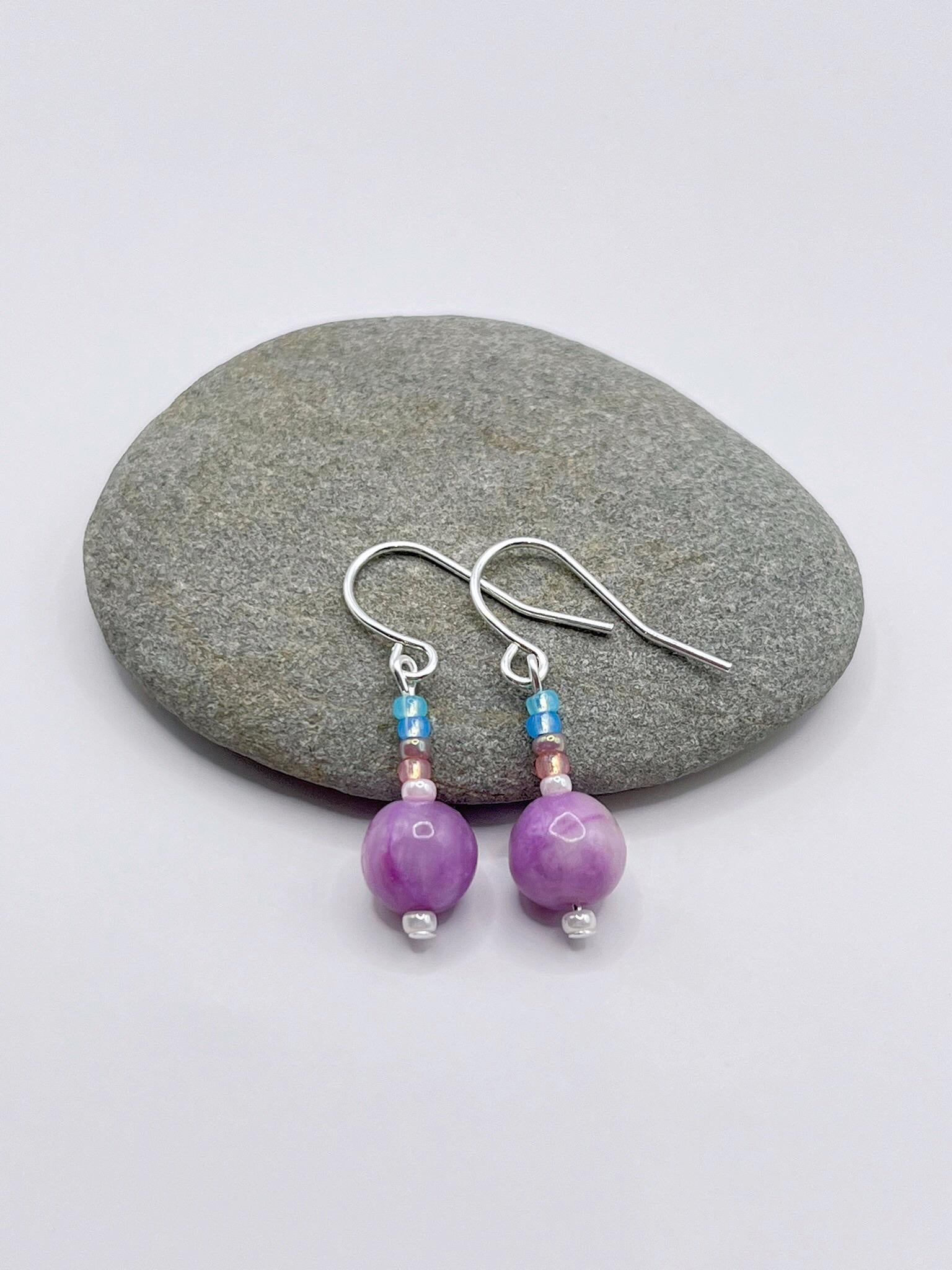 Pair of pastel seed bead and lilac jasper earrings laid on a grey pebble against a white background.
