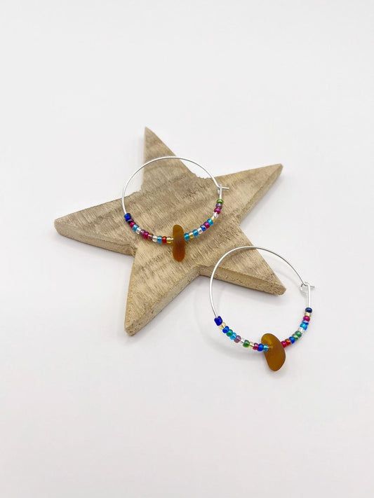 Rainbow seed bead and burnt orange sea glass hoop earrings laid on a wooden star against a white background