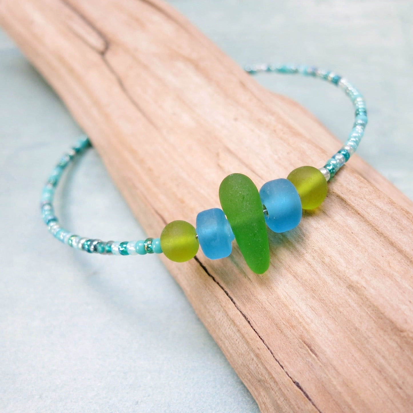 Close up of bracelet with green sea glass and turquoise and green glass beads laid on driftwood against a pale blue background