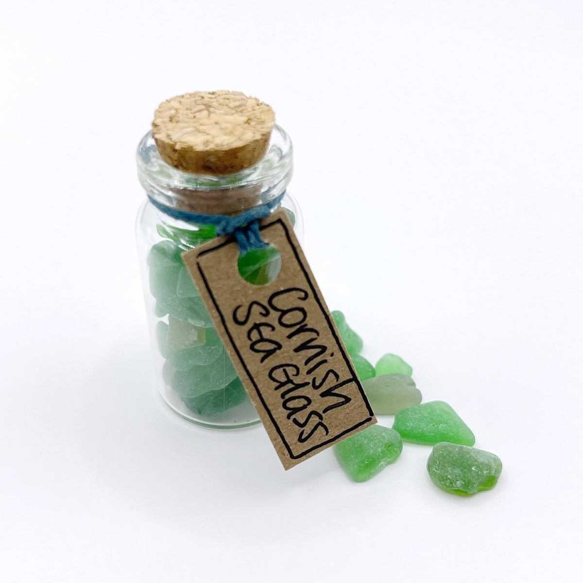 A small clear glass bottle with a cork stopper, filled with green sea glass and tied with a blue string. A brown paper tag hangs from the neck and reads 'CORNISH SEA GLASS.