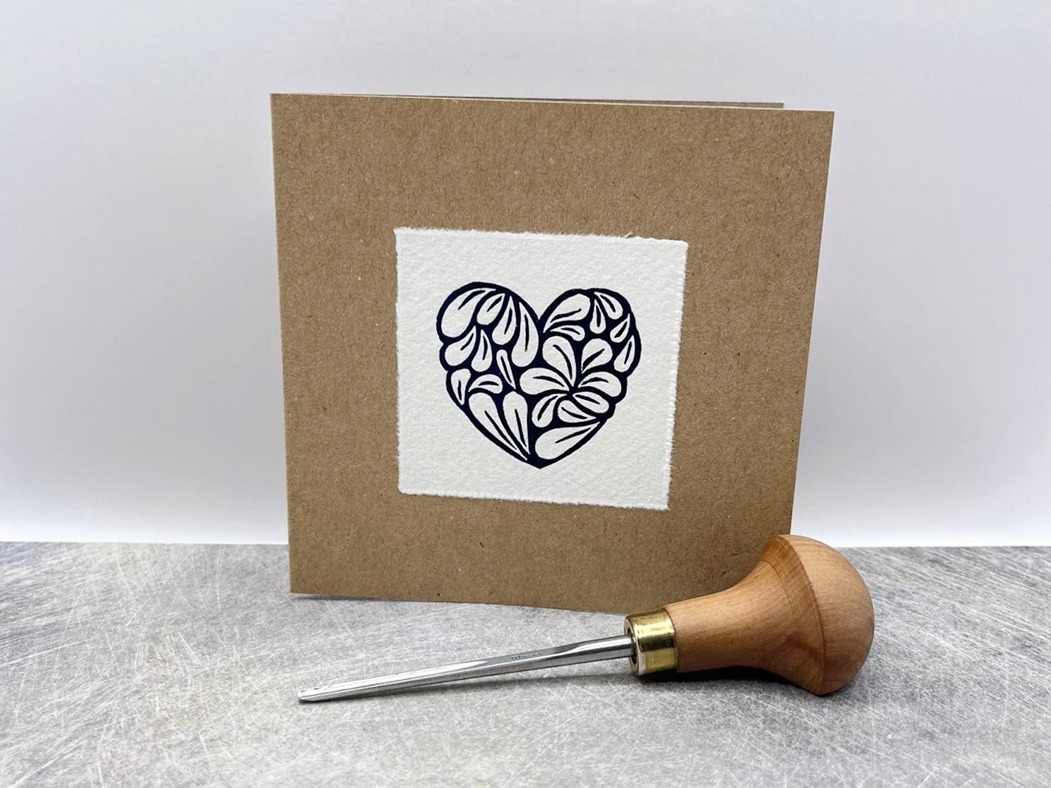 Kraft card with a white square featuring an original lino printed blue heart design stood against a white background. Photo includes lino cutting tool used.