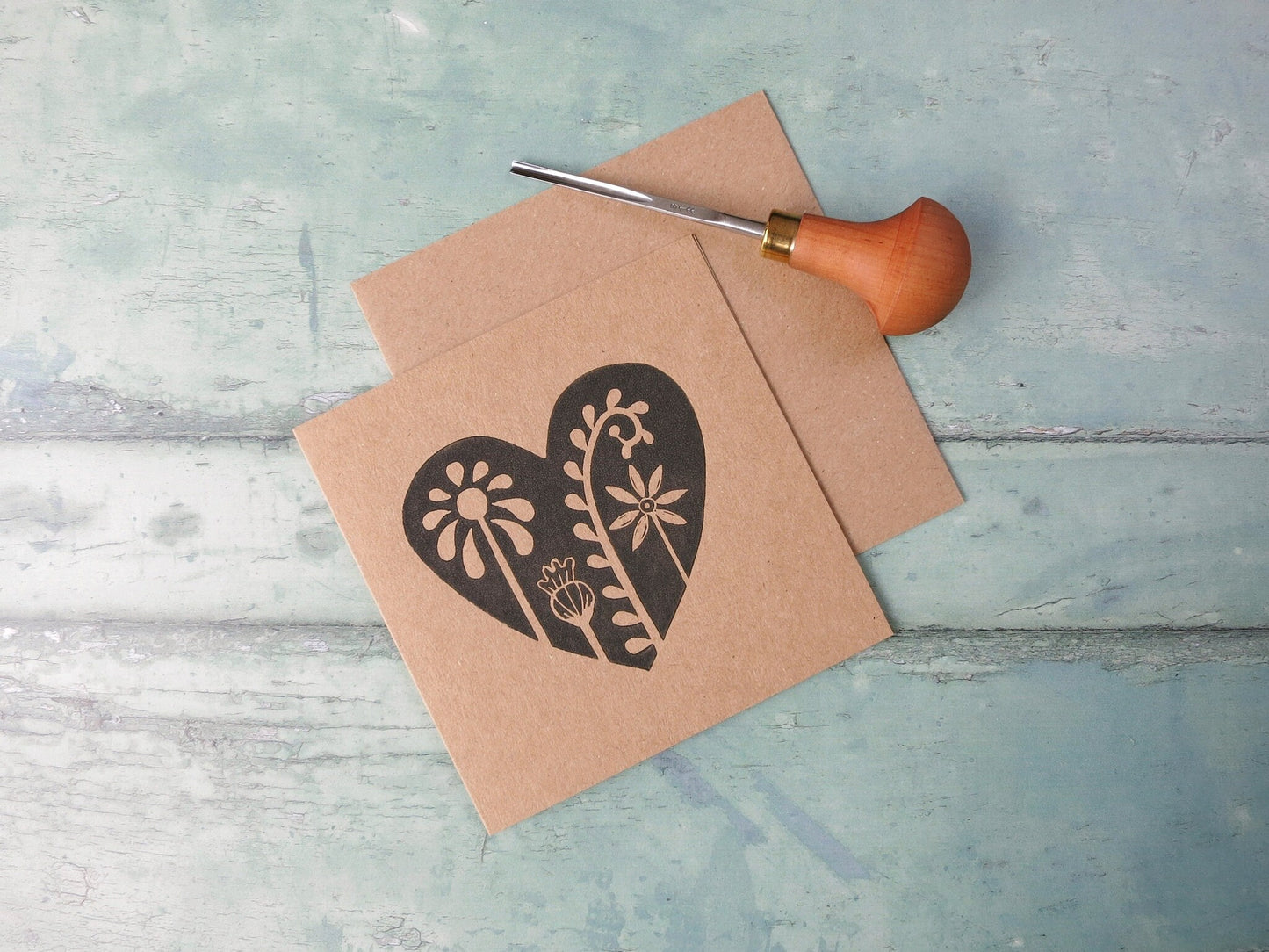 Kraft paper card with an original lino printed floral heart design laid on  a textured surface. Photo includes the lino cutting tool used.