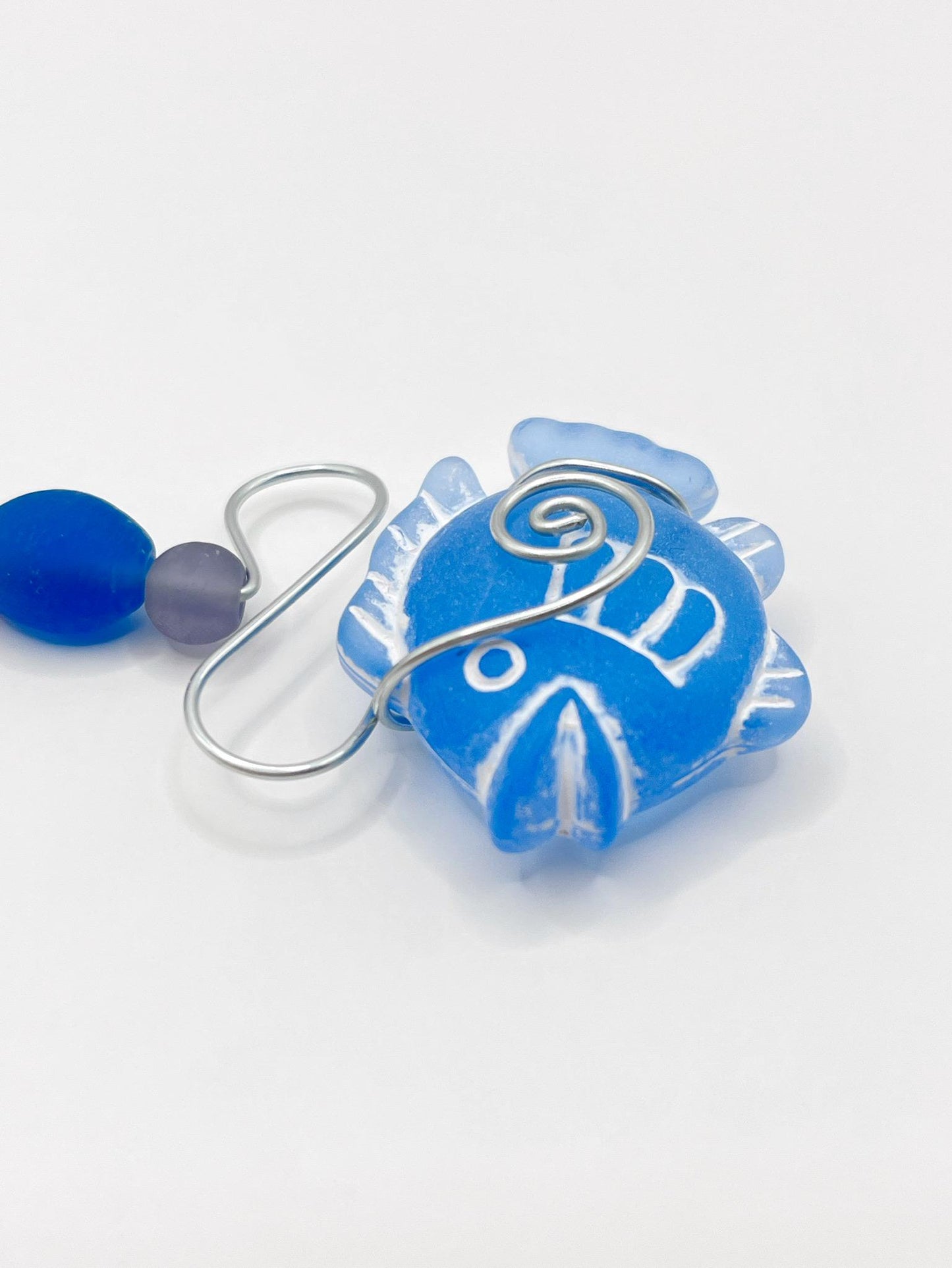 Close up of decorative hanging ornament with blue fish design, wire and colourful beads, laid  on a white background