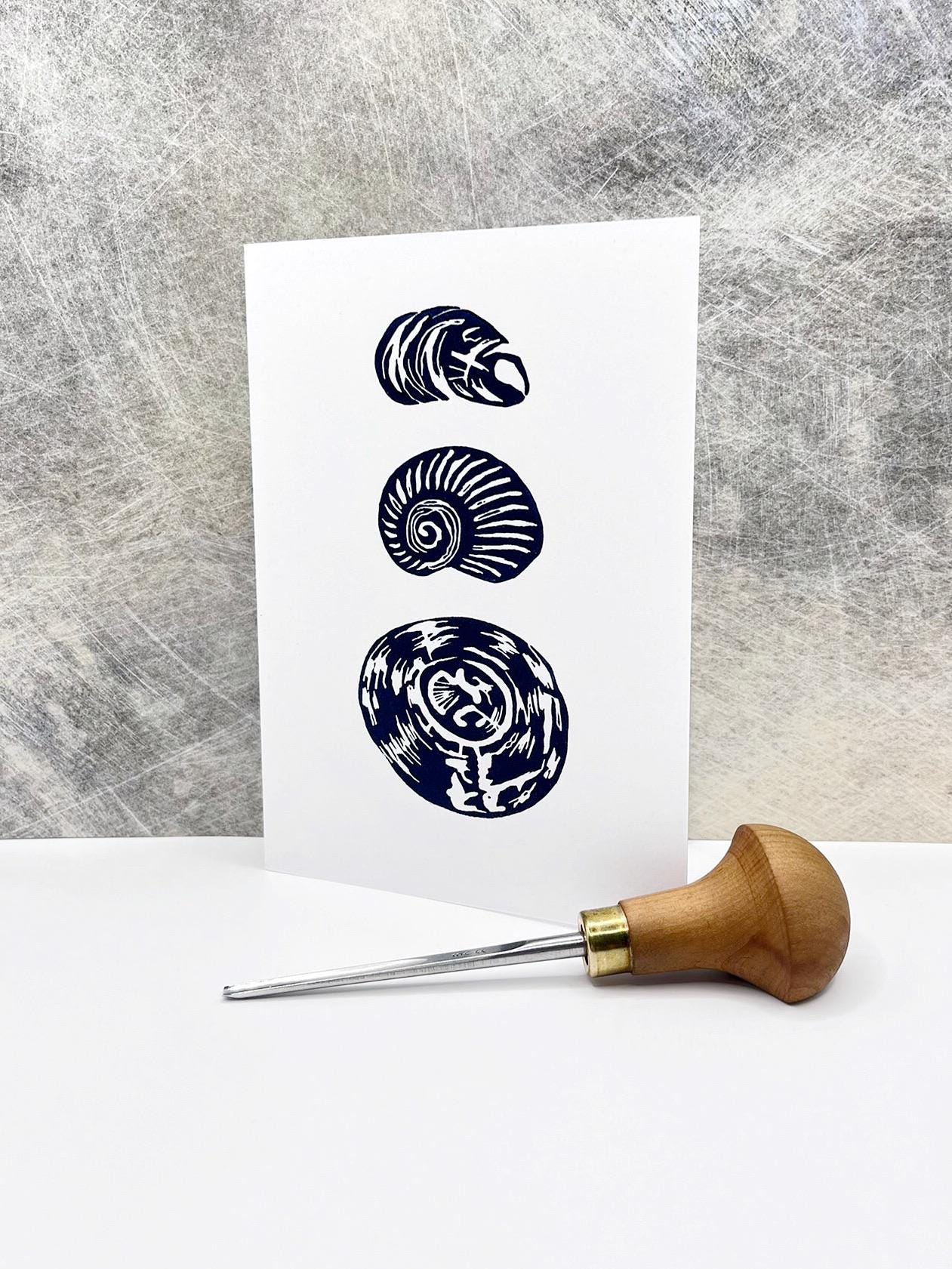 Three dark blue original lino print shells on a white card stood against a textured grey background. Included in the photo is the lino cutting tool used.