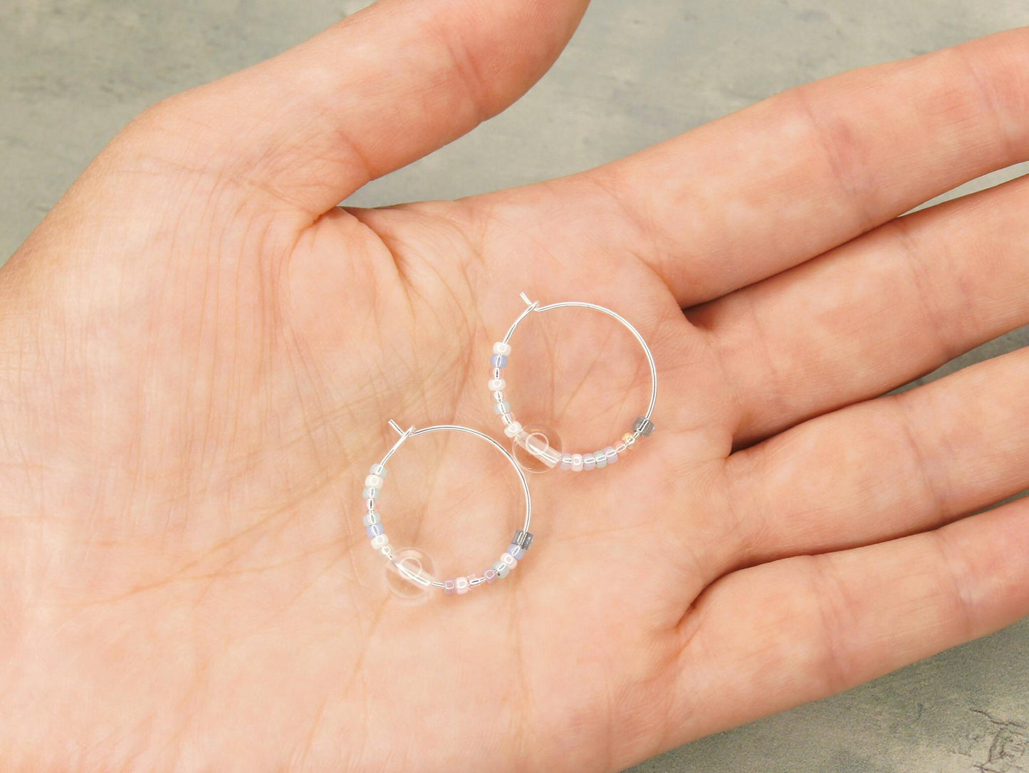 Pair of pastel seed bead and clear crystal hoop earrings held in a hand against a green background
