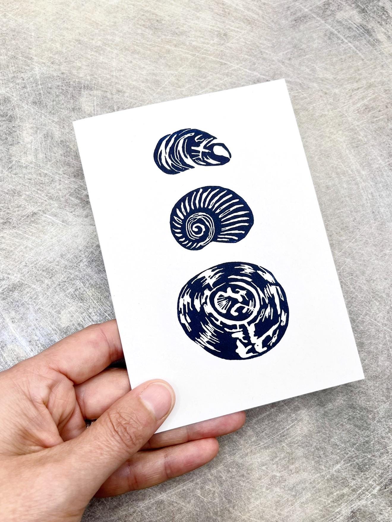 Hand holding a card with three blue shell lino prints held in my hand against a textured grey surface
