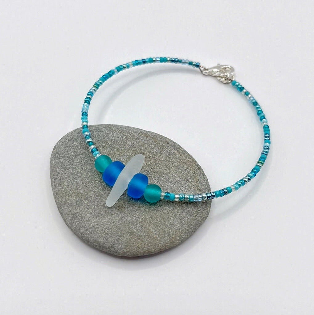 Bracelet with white sea glass and blue and turquoise beads laid on a grey pebble against a white background