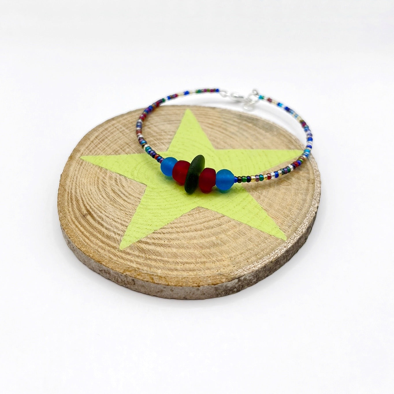 A rainbow coloured beaded bracelet with red and blue frosted glass beads and a piece of olive green coloured Cornish sea glass, finished with a silver plated lobster clasp and laid on a slice of wood with a pale lime star design.