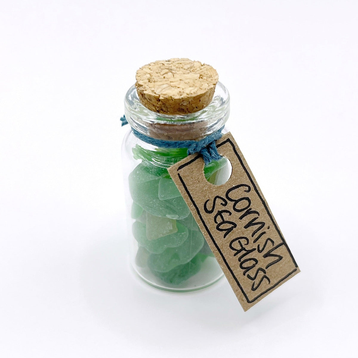 A small clear glass bottle with a cork stopper, filled with green sea glass pieces, tied with a blue string, and has a kraft paper tag that reads 'Cornish Sea Glass' on a white background.