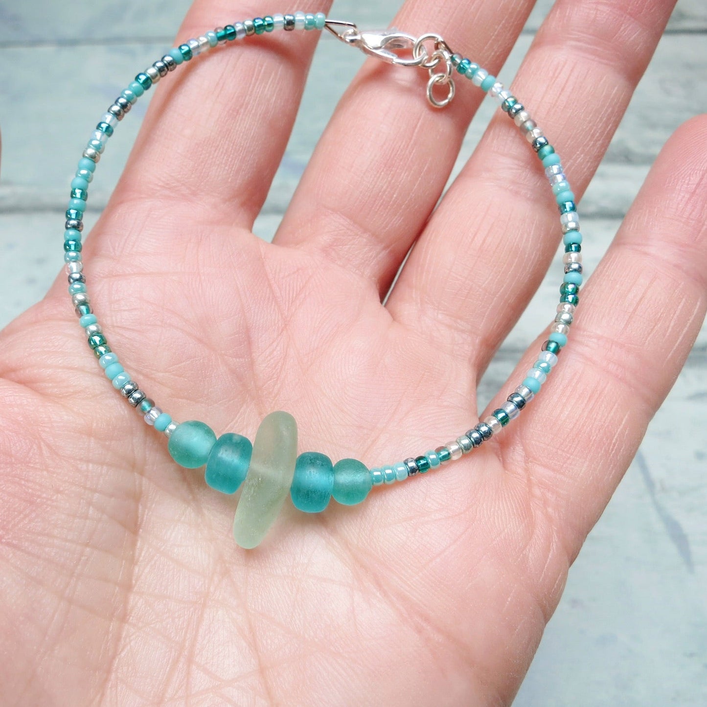 Bracelet with pale sea foam sea glass and sea green and turquoise glass beads held in a hand against a pale blue background