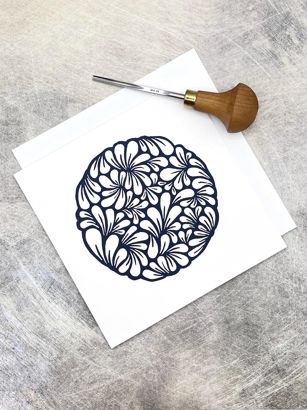 Original navy blue lino printed mussel shell pattern on white card laid on a textured background. Photo includes lino cutting tool.