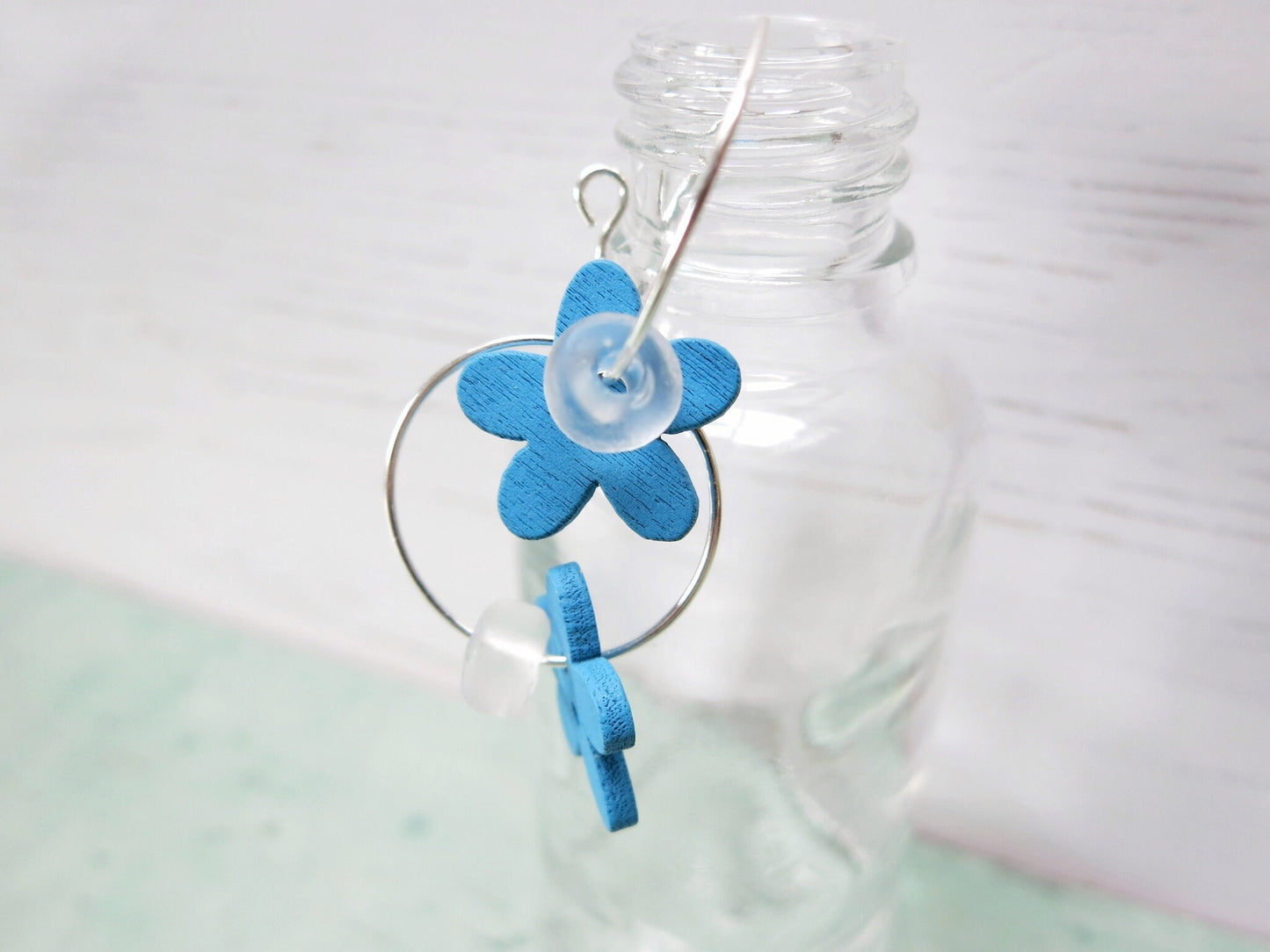 Close up of silver hoop earrings with Blue wooden flowers and a glass bead hung on a glass bottle.