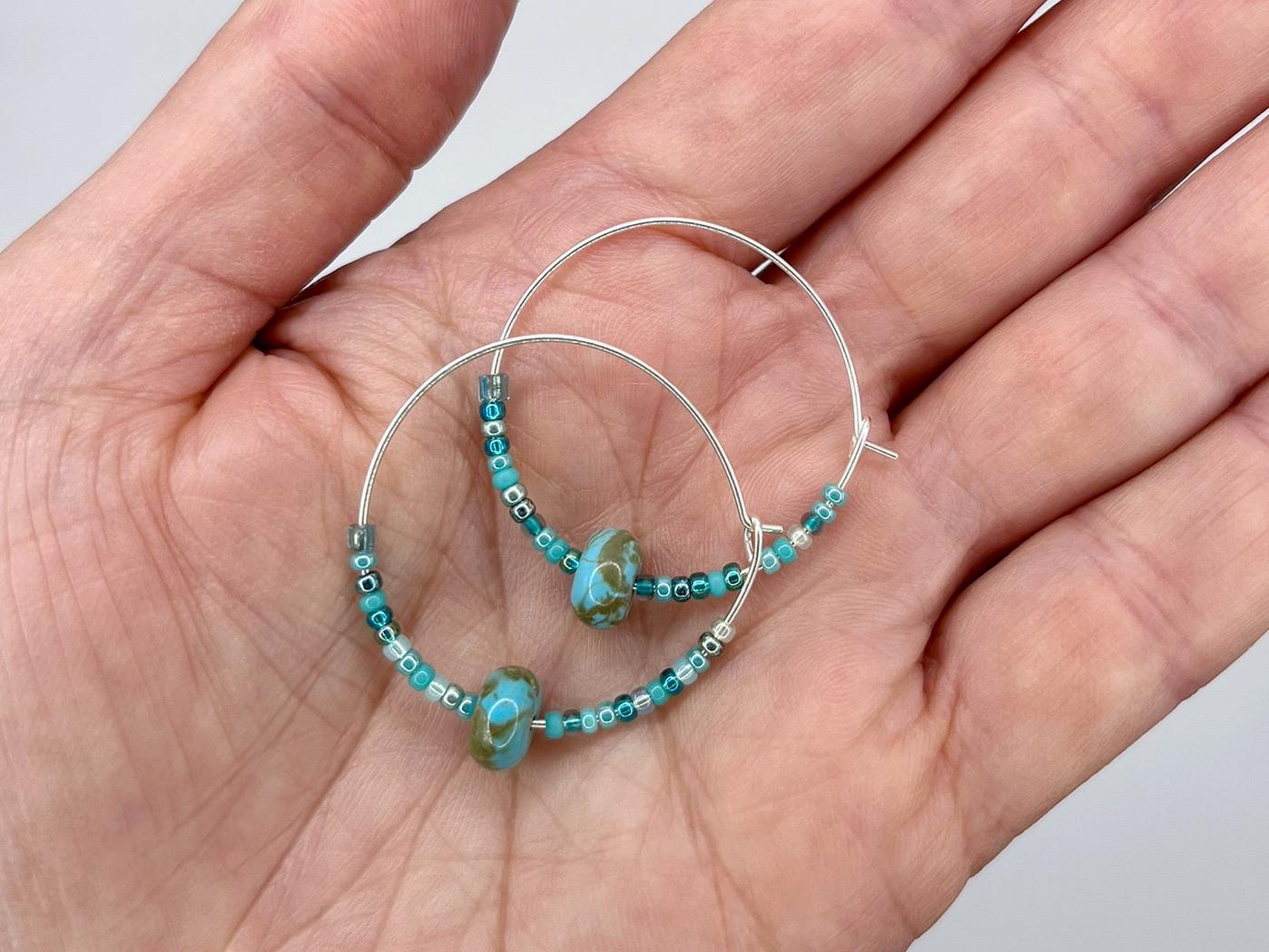Silver hoop earrings with turquoise beads held in a hand against a neutral background