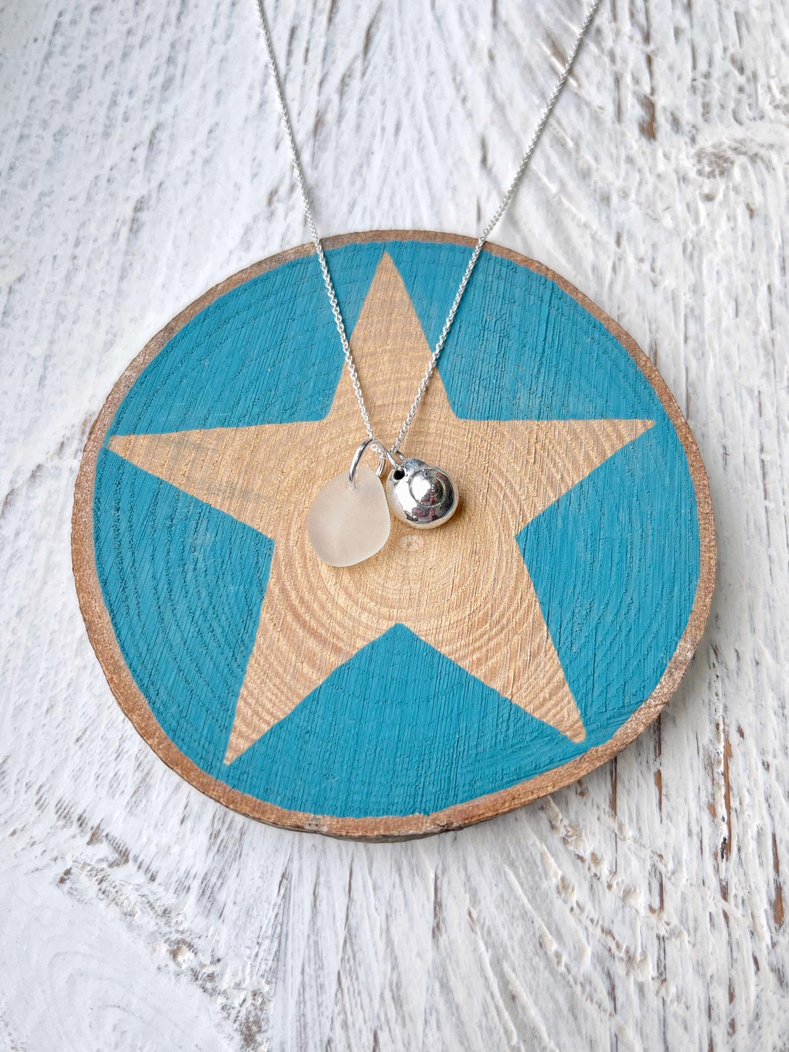 Necklace with a silver shell and white sea glass pendant laid on a star printed on wood sat  against a textured surface